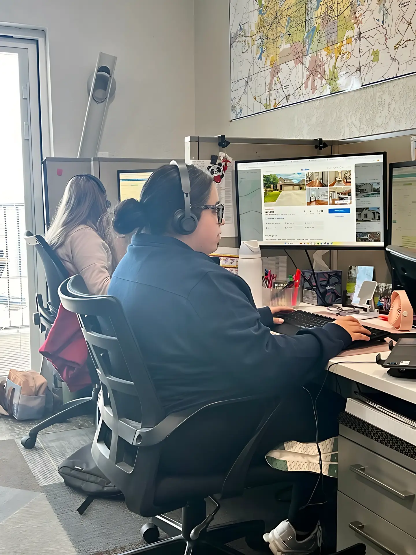 Two people working at computer workstations with headsets in an office, a large wall map on the wall behind them.