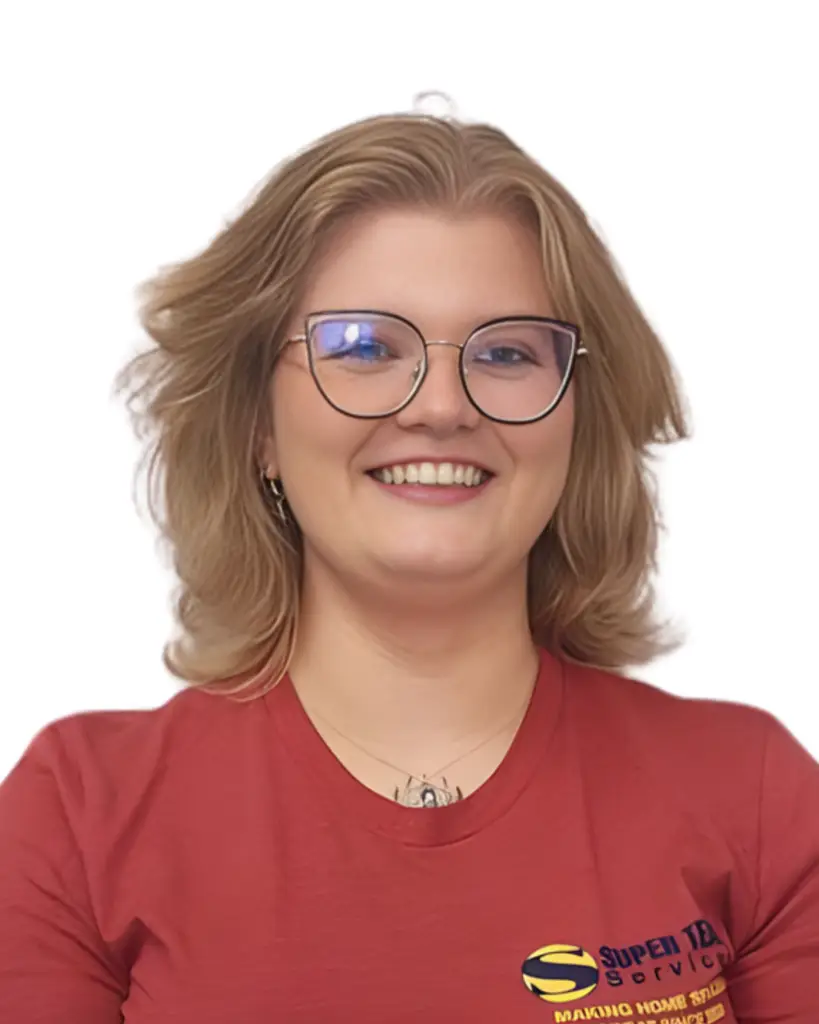 A person with shoulder-length blonde hair and round glasses smiles at the camera, wearing a red shirt with a yellow and blue logo on the chest, in front of a plain white background.