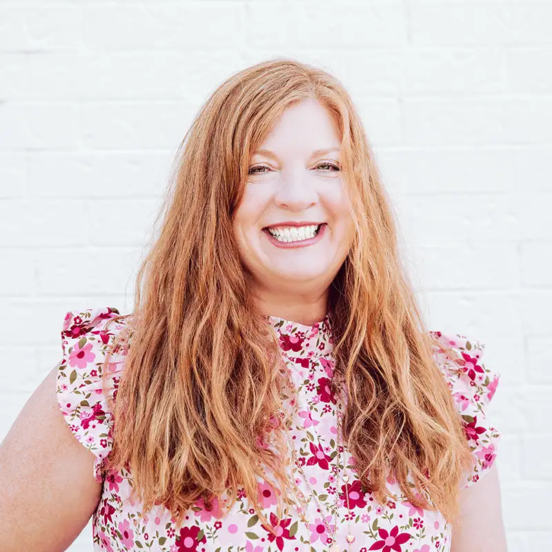 A woman with long, wavy, reddish-brown hair smiles broadly while standing in front of a white brick wall. She is wearing a floral top with pink, red, and white colors and ruffled sleeves.