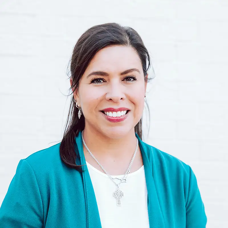 A woman with dark hair pulled back, wearing a teal blazer over a white top and silver jewelry, smiles in front of a light-colored brick wall.