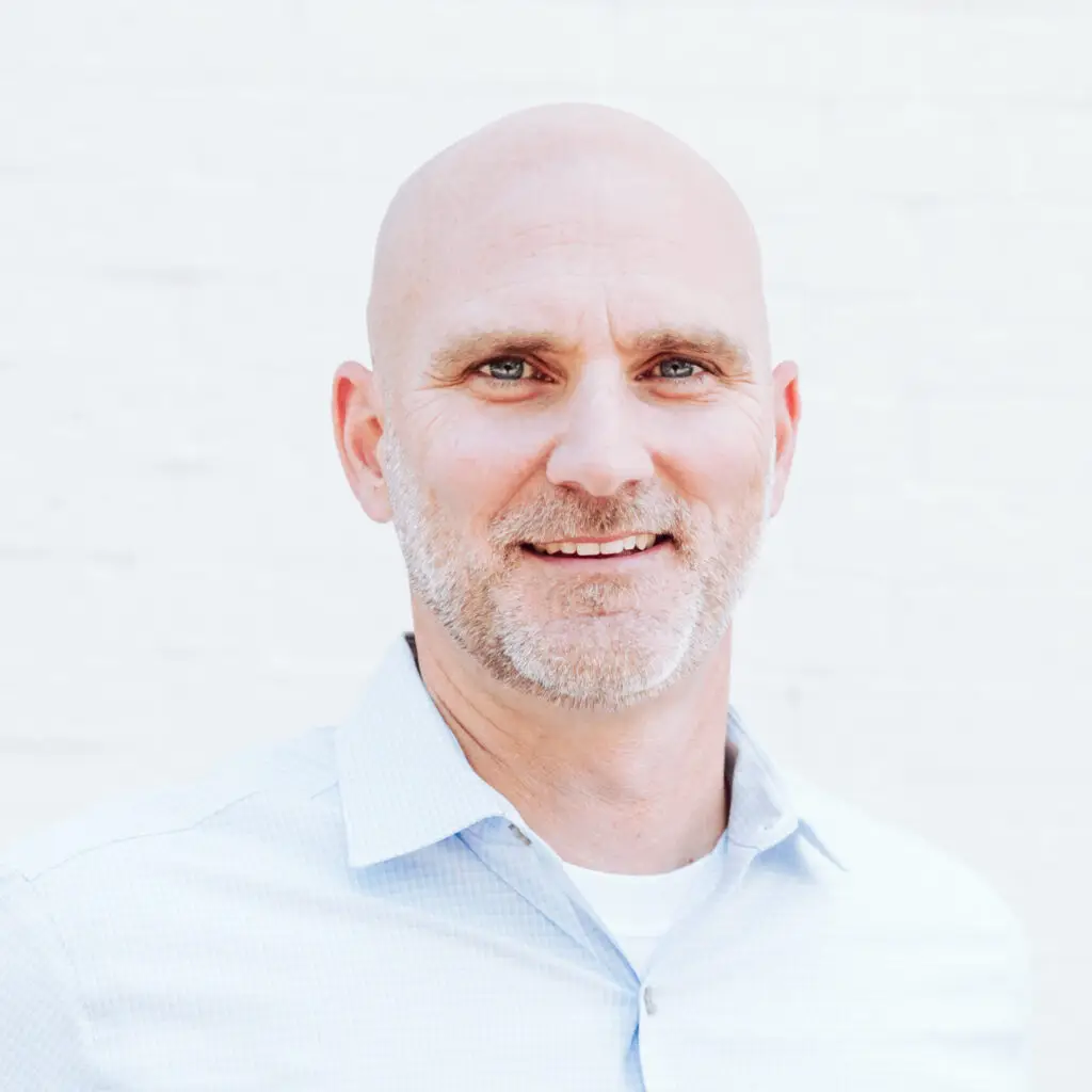 A middle-aged man with a shaved head and short beard is smiling. He is wearing a light blue collared shirt and standing in front of a white brick wall.