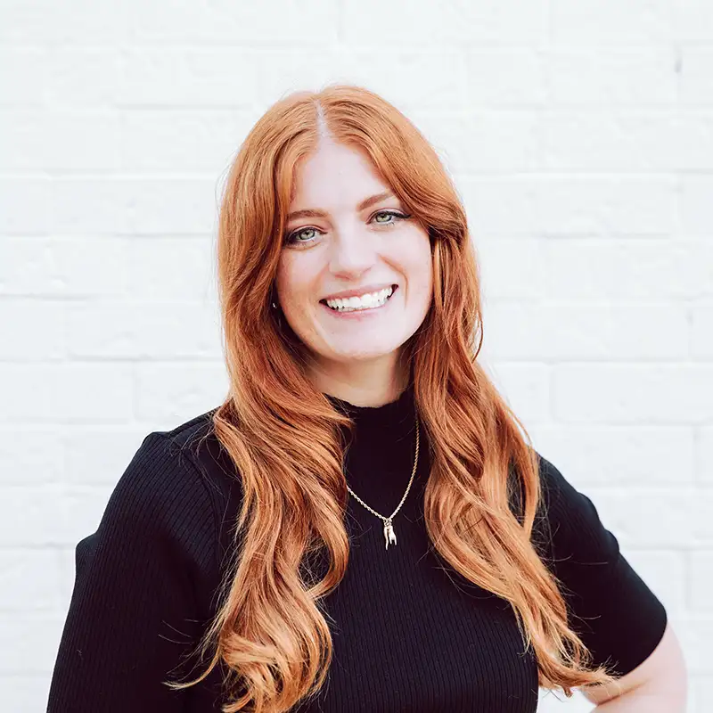 A woman with long red hair wearing a black top and a gold necklace stands smiling in front of a white brick wall.