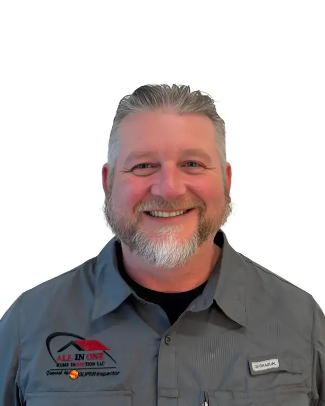 A middle-aged man with short gray hair and a beard, smiling, wearing a gray collared shirt with embroidered logos on the chest, standing against a plain white background.