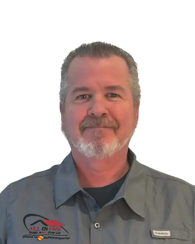 A middle-aged man with short gray hair and a beard, wearing a gray collared shirt with a company logo and name tag, smiles at the camera against a white background.